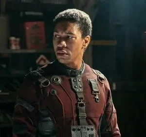 Um homem negro em uniforme futurista vermelho e preto olha tenso para o lado em cenário escuro e utilitário, cheio de objetos.