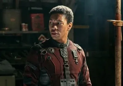Um homem negro em uniforme futurista vermelho e preto olha tenso para o lado em cenário escuro e utilitário, cheio de objetos.