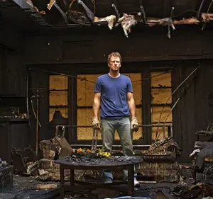 Homem de pé em uma sala de estar carbonizada, com móveis e teto danificados. Há flores coloridas em uma mesa central.