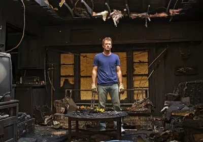 Homem de pé em uma sala de estar carbonizada, com móveis e teto danificados. Há flores coloridas em uma mesa central.
