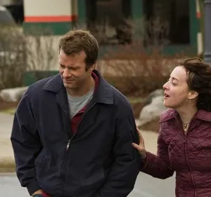 Homem de jaqueta azul olha para baixo, expressão sombria. Mulher de casaco vinho o toca e fala com urgência, em rua.