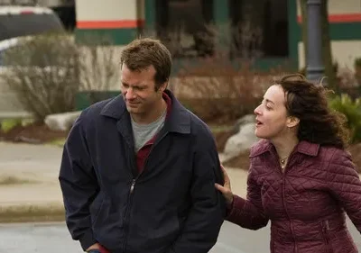 Homem de jaqueta azul olha para baixo, expressão sombria. Mulher de casaco vinho o toca e fala com urgência, em rua.