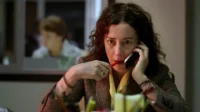 Mulher com cabelo cacheado e expressão intensa fala ao telefone, mordendo uma caneta. Fundo desfocado de escritório.