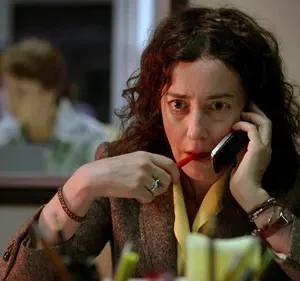 Mulher com cabelo cacheado e expressão intensa fala ao telefone, mordendo uma caneta. Fundo desfocado de escritório.