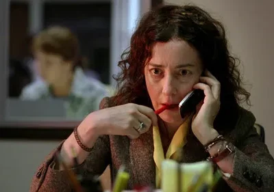 Mulher com cabelo cacheado e expressão intensa fala ao telefone, mordendo uma caneta. Fundo desfocado de escritório.