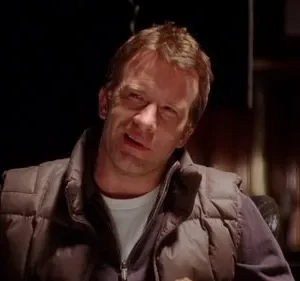 Pôster: Homem de meia-idade com colete marrom, expressão confusa e levemente cínica, em ambiente escuro e misterioso.