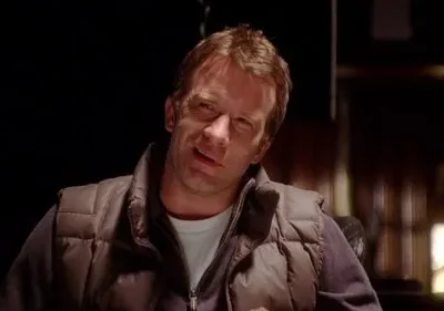 Pôster: Homem de meia-idade com colete marrom, expressão confusa e levemente cínica, em ambiente escuro e misterioso.