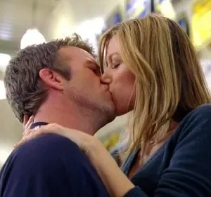 Um homem e uma mulher loira se beijam apaixonadamente. O fundo desfocado revela um local público com luzes fluorescentes.