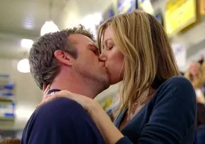 Um homem e uma mulher loira se beijam apaixonadamente. O fundo desfocado revela um local público com luzes fluorescentes.