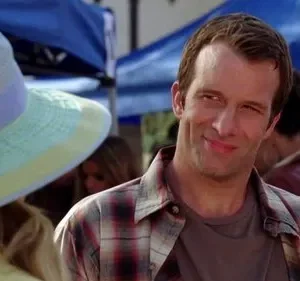 Homem com camisa xadrez e sorriso maroto. Chapéu colorido à esquerda. Cena externa diurna com outras pessoas.