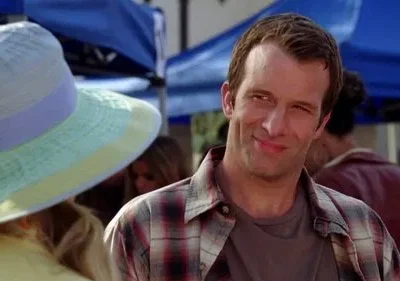 Homem com camisa xadrez e sorriso maroto. Chapéu colorido à esquerda. Cena externa diurna com outras pessoas.