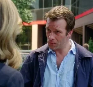 Homem de jaqueta azul e camisa aberta, com expressão séria, olhando para uma mulher loira de costas. Fundo urbano borrado.