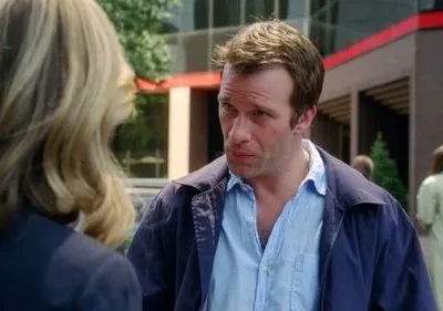 Homem de jaqueta azul e camisa aberta, com expressão séria, olhando para uma mulher loira de costas. Fundo urbano borrado.