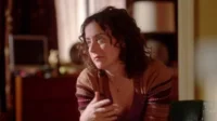 Mulher de meia-idade com cabelo encaracolado e cardigã marrom, parece pensativa. Ambiente interno com iluminação suave, fundo escuro com móveis desfocados.