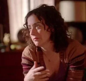 Mulher de meia-idade com cabelo encaracolado e cardigã marrom, parece pensativa. Ambiente interno com iluminação suave, fundo escuro com móveis desfocados.