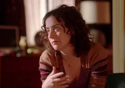 Mulher de meia-idade com cabelo encaracolado e cardigã marrom, parece pensativa. Ambiente interno com iluminação suave, fundo escuro com móveis desfocados.