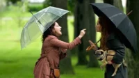 Duas mulheres com guarda-chuvas discutem num parque. Uma ruiva segura um pug, a outra gesticula em fúria sob chuva leve.