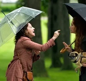 Duas mulheres com guarda-chuvas discutem num parque. Uma ruiva segura um pug, a outra gesticula em fúria sob chuva leve.