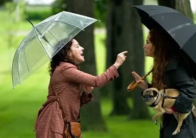 Duas mulheres com guarda-chuvas discutem num parque. Uma ruiva segura um pug, a outra gesticula em fúria sob chuva leve.