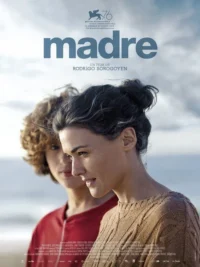 Duas mulheres na praia. Uma,de suéter marrom,sorri. Outra,com cabelo cacheado e camisa vermelha,desfocada ao fundo. Céu nublado.