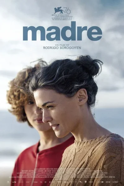 Duas mulheres na praia. Uma, de suéter marrom, sorri. Outra, com cabelo cacheado e camisa vermelha, desfocada ao fundo. Céu nublado.