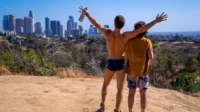 Dois homens em um morro ensolarado com vista para uma cidade. Um, sem camisa, ergue os braços em celebração alegre.