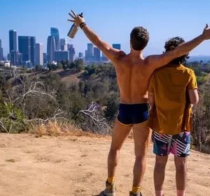 Dois homens em um morro ensolarado com vista para uma cidade. Um, sem camisa, ergue os braços em celebração alegre.