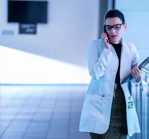 Mulher de jaleco, óculos e pasta, fala ao telefone em corredor iluminado em tons de azul. Ambiente profissional e sério.