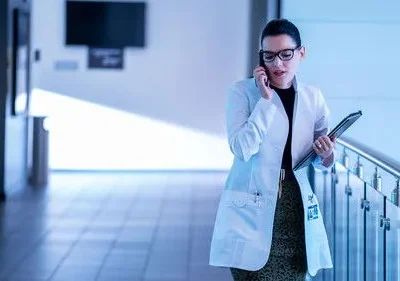 Mulher de jaleco, óculos e pasta, fala ao telefone em corredor iluminado em tons de azul. Ambiente profissional e sério.