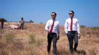 Dois homens de camisa branca e gravata vermelha andam em campo seco. Ao fundo, um animal morto e pessoas. Clima misterioso.