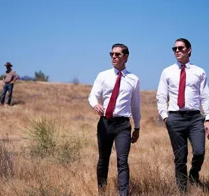 Dois homens de camisa branca e gravata vermelha andam em campo seco. Ao fundo, um animal morto e pessoas. Clima misterioso.