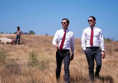 Dois homens de camisa branca e gravata vermelha andam em campo seco. Ao fundo, um animal morto e pessoas. Clima misterioso.