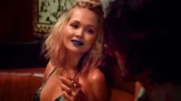 Mulher loira de batom azul e top brilhante segura coquetel, olhando para um homem em um bar escuro. Ambiente íntimo e misterioso.