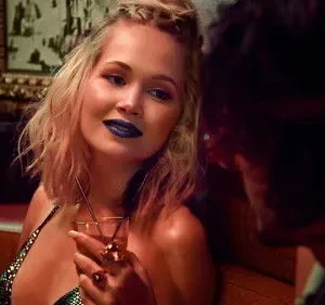 Mulher loira de batom azul e top brilhante segura coquetel, olhando para um homem em um bar escuro. Ambiente íntimo e misterioso.