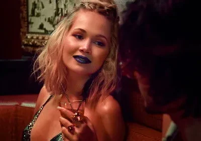 Mulher loira de batom azul e top brilhante segura coquetel, olhando para um homem em um bar escuro. Ambiente íntimo e misterioso.