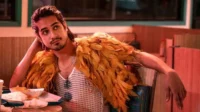 Jovem de cabelo escuro e barba usa regata de malha e estola de penas laranja, pensativo em um restaurante com luz avermelhada.