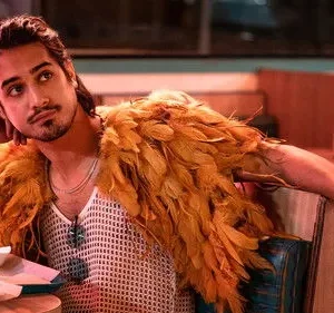 Jovem de cabelo escuro e barba usa regata de malha e estola de penas laranja, pensativo em um restaurante com luz avermelhada.