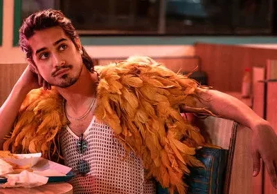 Jovem de cabelo escuro e barba usa regata de malha e estola de penas laranja, pensativo em um restaurante com luz avermelhada.