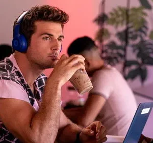 Jovem com fones azuis e camisa estampada bebe café, olhando pensativo. Cenário moderno com laptop e luz roxa.