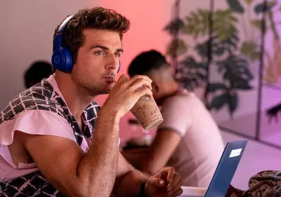 Jovem com fones azuis e camisa estampada bebe café, olhando pensativo. Cenário moderno com laptop e luz roxa.