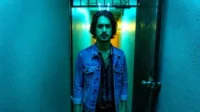 Homem de jaqueta jeans olha intenso em corredor azul-neon. Luz amarela no fundo cria clima sombrio.