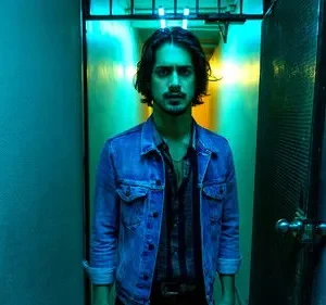 Homem de jaqueta jeans olha intenso em corredor azul-neon. Luz amarela no fundo cria clima sombrio.