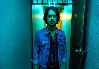 Homem de jaqueta jeans olha intenso em corredor azul-neon. Luz amarela no fundo cria clima sombrio.