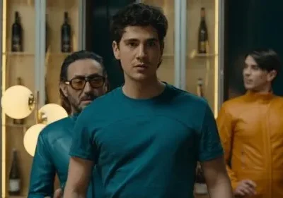 Jovem de camisa azul-petróleo, olhos fixos. Homem de óculos escuros e outro de jaqueta laranja em cenário moderno e estilizado. Clima tenso.