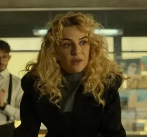 Mulher loira atenta em primeiro plano, homem de óculos e suspensórios atrás. Cenário interno com luz dramática e tensa.