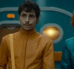 Homem de bigode com jaqueta laranja e mulher ruiva de jaqueta azul. Ambos sérios, em cenário futurista azul-petróleo com luzes.