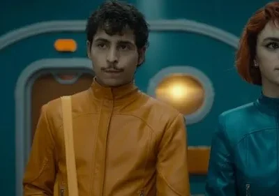 Homem de bigode com jaqueta laranja e mulher ruiva de jaqueta azul. Ambos sérios, em cenário futurista azul-petróleo com luzes.