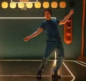 Um jovem dança em quadra de basquete estilizada com luzes neon e paredes verdes. Bolas laranjas e assento ao fundo. Cena vibrante e moderna.