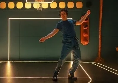 Um jovem dança em quadra de basquete estilizada com luzes neon e paredes verdes. Bolas laranjas e assento ao fundo. Cena vibrante e moderna.