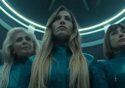 Um grupo de mulheres com roupas azuis-petróleo e expressão séria olha para cima num ambiente futurista com anéis de luz neon.